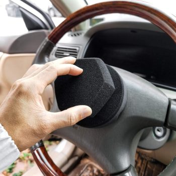 Person placing a black foam steering wheel cover over a car steering wheel with wood trim, viewed from the driver's side.